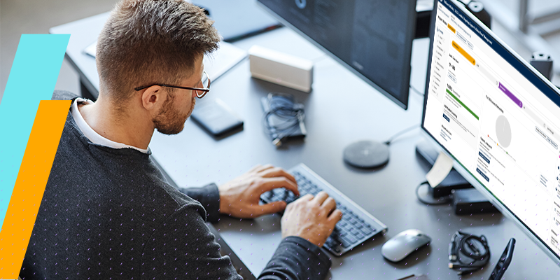 Man wearing glasses typing on a compact keyboard at a dual-monitor desk setup