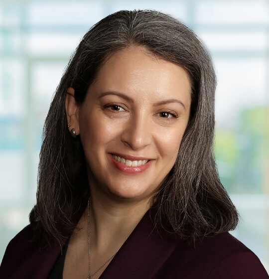 Professional woman smiling at camera with shoulder-length gray hair, wearing a burgundy blazer in an office setting, head-and-shoulders portrait.